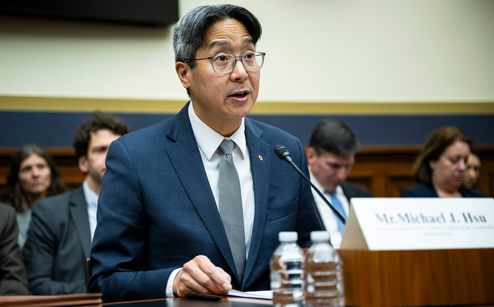 Acting Comptroller of the Currency Michael Hsu, wearing a dark suit, gray tie, and glasses, speaks into a microphone at a hearing. He holds a paper in his hand, with water bottles and a nameplate visible in front of him. Other attendees are seated in the background, blurred.